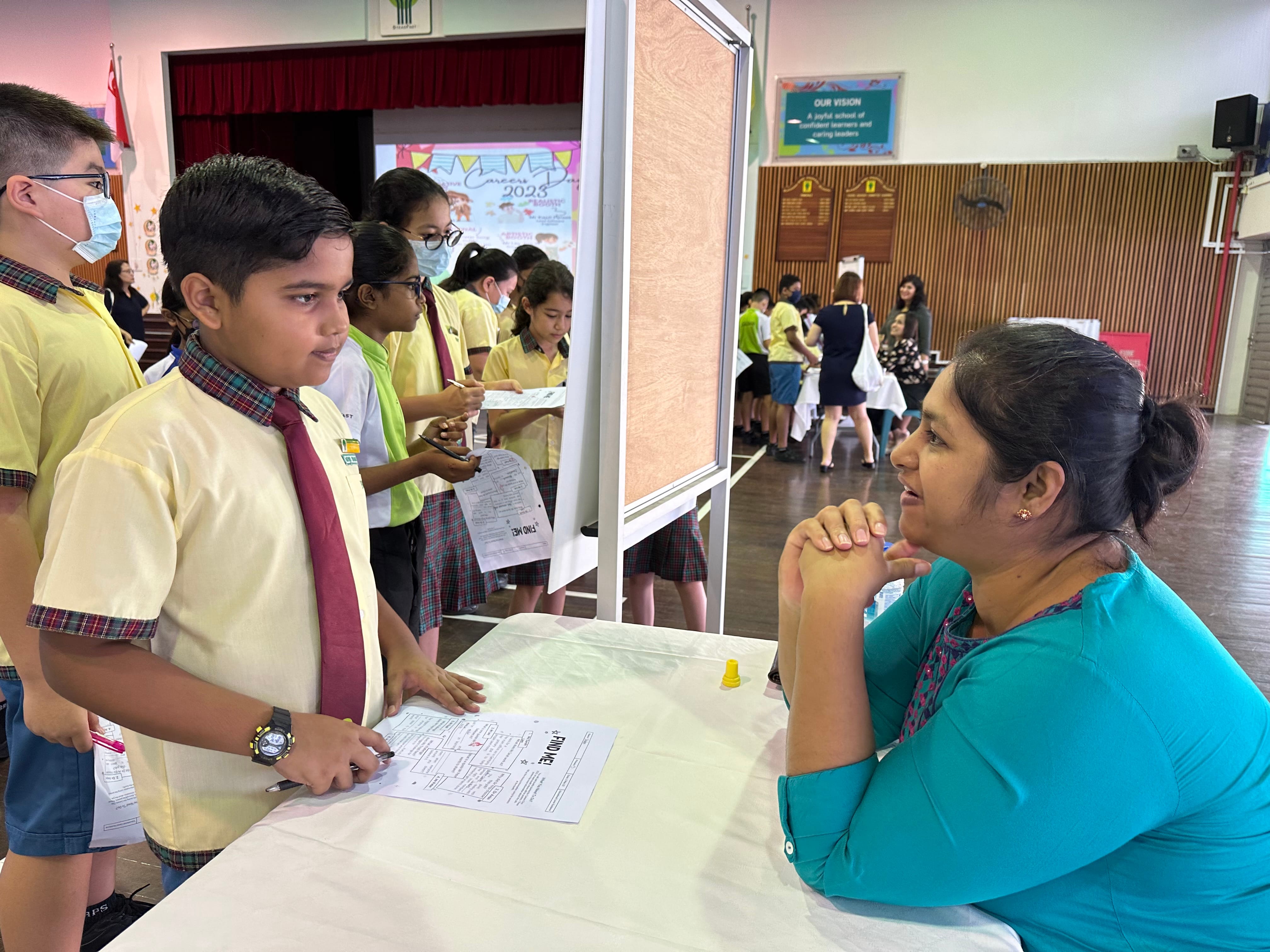 A school boy in yellow uniform and red tie speaks with a woman in teal across a table in a school hall.